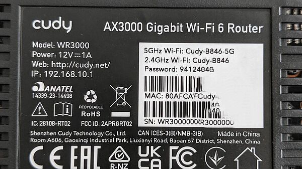 Rückseite des Cudy WR3000 mit Typenschild. Das Etikett zeigt Modellbezeichnung, Stromversorgung (12V 1A), Seriennummer, MAC-Adresse, WLAN-SSID für 2,4 GHz und 5 GHz sowie Herstellerinformationen und Zertifizierungslogos (CE, FCC, RoHS u.a.).